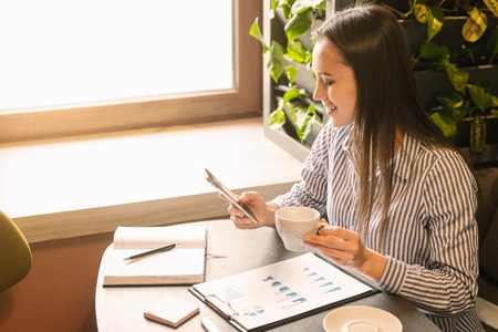 Young accountant making financial report in cafe, drinking coffee and using cellphone, empty spaceの写真素材