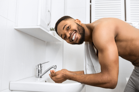 Smiling afro man with bare muscular torso washing face in bathroom, looking at camera Morning hygiene concept, copy spaceの写真素材