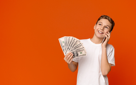 Cheerful teenage boy holding money banknotes and talking on mobile phone, orange panorama background with empty spaceの写真素材
