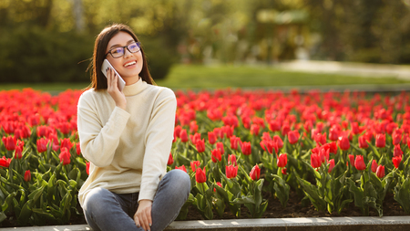 Happy Girl Talking On Phone Near Red Tulips In Spring Park, panorama. free spaceの写真素材
