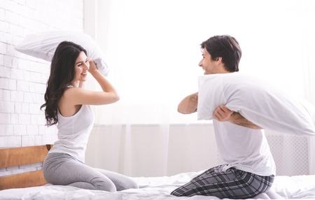 Cheerful couple in love is fighting by pillows in bed, having fun together at home, free spaceの写真素材