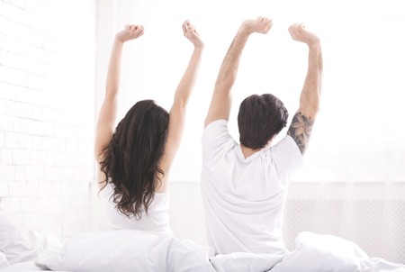 Well rested young couple waking up in morning, stretching on bed together, back view, free spaceの写真素材