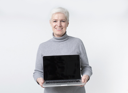 Modern technology and senior age. Cheerful mature woman showing blank screen on laptop computer, light studio background, empty spaceの写真素材