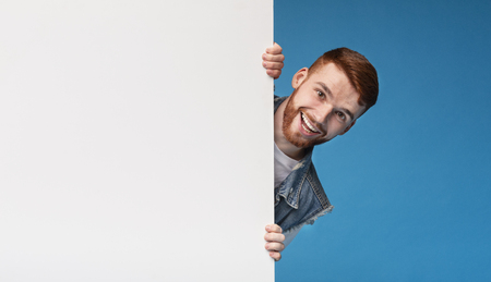 Cheerful young smiling guy looking out of white blank placard ready for text, information or product, blue backgroundの写真素材