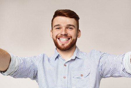 Selfie everywhere! Handsome young caucasian man photographing himself on camera and widely smiling, standing against beige background