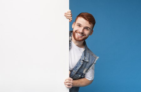 Happy smiling guy looking out of white blank placard ready for advertisement, blue backgroundの写真素材