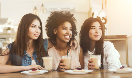 Three smiling multiracial girls sitting in cafe looking aside, drinking coffee and having enjoyable timeの写真素材