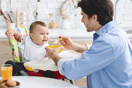 Dad feeding his baby with healthy food, grimacing infant doesnt like taste, free space