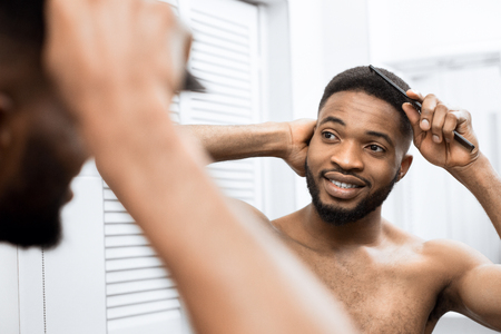 Smiling afro man with naked torso combing hair and looking in mirror in bathroom.の写真素材