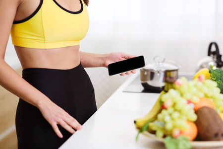 Calorie Counter App. Sporty Girl Using Phone At Kitchen, Fruits On Tableの写真素材