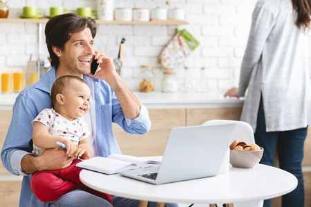 Father Working From Home With Baby Son. Talking on Phone and Using Laptop in Kitchen, Free Spaceの写真素材