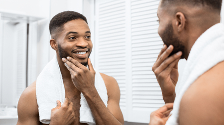 Happy afro man with bare torso touching beard looking in mirror at bathroom. Modern man in bathroom conceptの写真素材