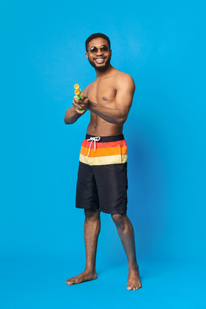 Get Ready To Be Wet! African american guy in swimwear shooting at camera with water gun, blue backgroundの写真素材