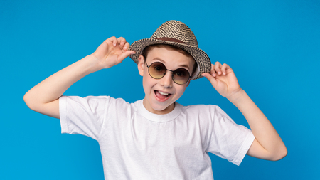 Portrait of cheerful stylish boy in sunglasses and wicker hat, blue panorama backgroundの写真素材