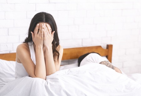 Relationship problems. Upset woman sitting on bed and crying, her fiance is sleeping, empty spaceの写真素材
