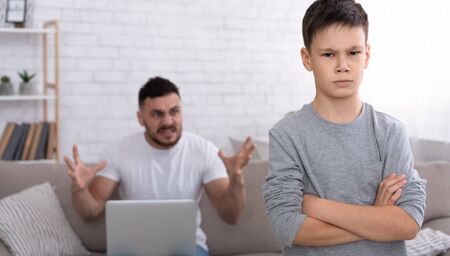 Generations misunderstanding. Sad little boy looking at camera with crossed hands, angry father with laptop yelling at him in background, free spaceの写真素材