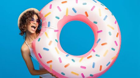 Hunger at the beach. Cheerful african girl biting her inflatable circle shaped like donut, blue...