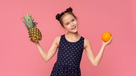 Natural summer nutrition. Cheerful little girl holding fresh pineapple and orange fruit on pink studio backgroundの写真素材