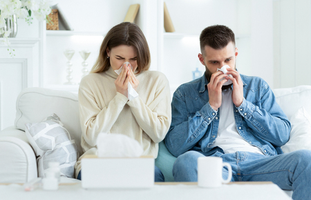 Couple Suffering From Cold, Blowing Noses And Sneezing, Sitting On Sofa At Homeの写真素材