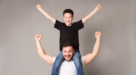 Excited man and boy enjoying success together, dad holding his son on shoulders, gray panorama backgroundの写真素材