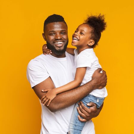 Happy african man embracing his positive little daughter, orange studio background