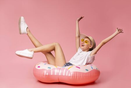 Positive millennial blonde sitting on big inflatable ring, ready to swim, pink studio backgroundの写真素材