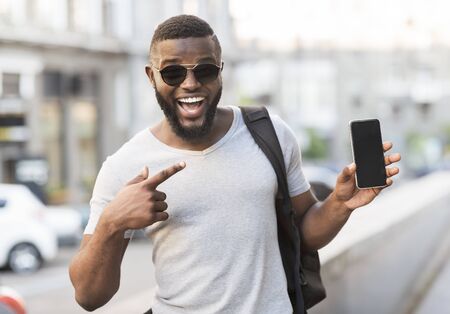 Hey something new. Happy african guy pointing his cellphone with black blank screen for advertisement outdoor, panoramaの写真素材