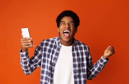 Amazing News. African Guy Holding Smartphone and Celebrating Success, Orange Backgroundの写真素材