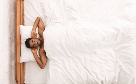 Naked african-american guy sleeping in bed with hands behind head, top view, free spaceの写真素材