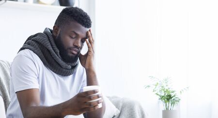 Sad african-american man with flu wrapped in warm scarf, holding cup with hot tea, free spaceの写真素材