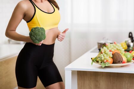 Weight loss concept. Girl holding broccoli and showing thumb up, cooking healthy salad in kitchenの写真素材