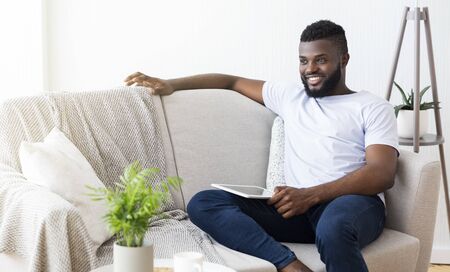 Lazy weekend at home. Cheerful black man sitting on couch with digital tablet, free spaceの写真素材