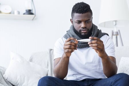 Ill african american man covered with scarf holding thermometer, having fever, free spaceの写真素材