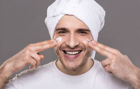 Morning hygiene. Millennial guy with towel on his head, applying cream on face, smiling at cameraの写真素材