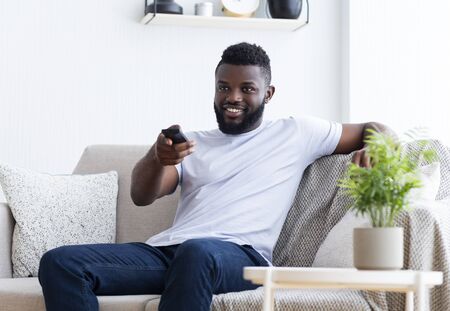 Watching TV at home. Young african american man having rest, switching channels with remote...