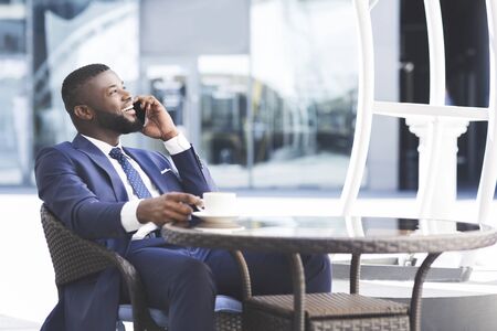 Cheerful Afro Businessman Having Coffee And Talking On Mobile Phone In Cafe. Selective focus. Panorama With Copy Spaceの写真素材