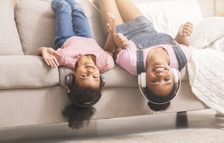 Teen and little black girls enjoying music in headphones, lying on couch headfirst looking at camera at homeの写真素材