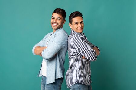Male Friendship. Two Smiling Arab Men Standing Back-To-Back Over Turquoise Studio Background. Panorama, Copy Spaceの写真素材