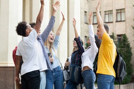 Happy students standing near university with raised hands, ready for exams