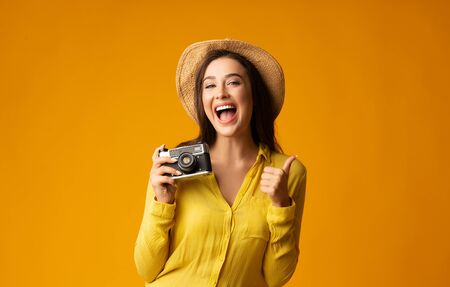 Excited Traveler Girl With Retro Camera Gesturing Thumb Up Over Yellow Studio Background. Free Space