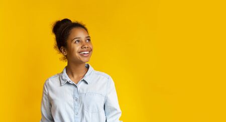 Young pensive african girl looking upwards at empty space on yellow studio background, panorama