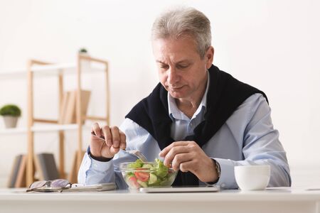 Senior businessman eating fresh vegetable salad, having break, working at home officeの写真素材