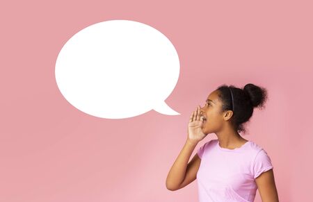 Beautiful African American girl making announcement at white blank speech bubble standing against pink background. Side view with copy spaceの写真素材