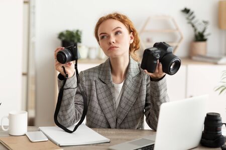 Photographer choosing photocamera for shooting, having doubts, working in office on laptopの写真素材