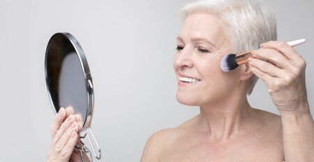 Portrait of beautiful senior woman doing makeup, applying blush to her cheek, panoramaの写真素材