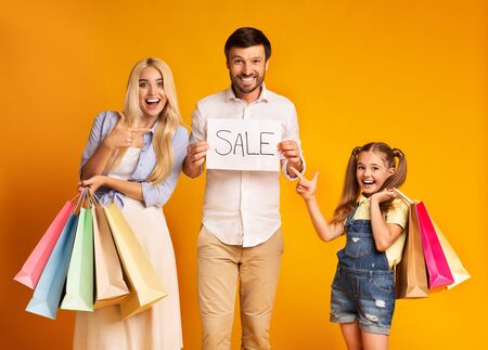 Family Shopping. Parents And Daughter Pointing Fingers At Sale Sign Holding Shopper Bags On Yellow Studio Background.の写真素材