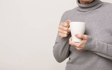 Closeup of female hands with coffee cup. Unrecognizable senior woman drinking hot beverage indoors,...