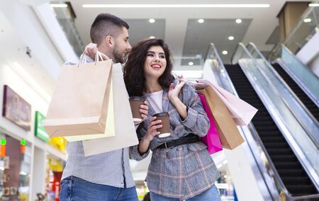 Happy millennial couple resting in mall, drinking coffee and walking after shopping