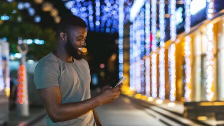 Smiling young african american man holding latest cellphone, chatting with friends, walking by night city, copy spaceの写真素材