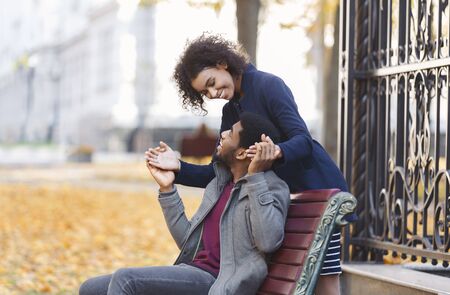 Peek a boo. Black young woman standing behind her man, guy holding her hands, sitting at park bench,...
